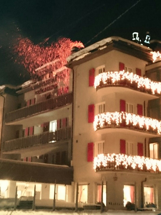 Kaminbrand eines Wohnhauses oder Hotels. Man sieht von Aussen wie der Kamin brennt und zwei Feuerwehrmänner den Brand (Funken sprühen) löschen wollen - Peter Leuenberger AG in Attiswil (Region Solothurn, Bern und Aargau)